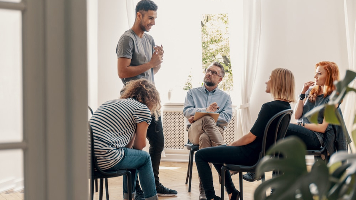 Adults sitting in a circle with one standing. Inpatient rehab programs provide structured support and customized treatment