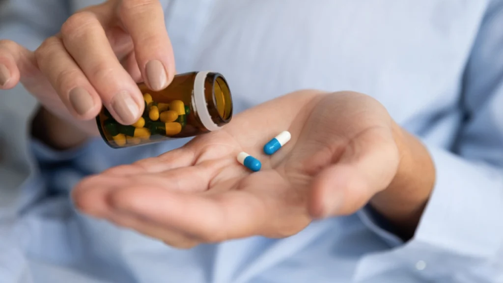 Hand holding a pill bottle and shaking blue and white pills into another hand. Medications are one treatment option for alcoholism.